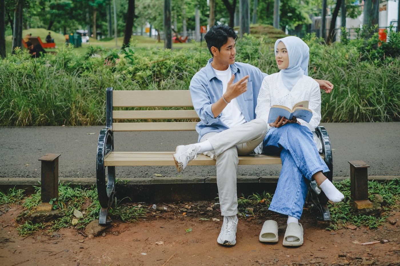 야외 공원 벤치에 나란히 앉아 휴대폰을 보며 웃고 있는 두 사람, 주변에는 잔잔한 조명과 푸른 잔디가 펼쳐져 있는 여유로운 저녁 데이트 장면